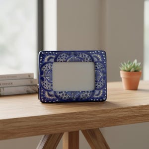 May include: A rectangular picture frame featuring a vibrant blue and white hand-painted pattern with intricate floral and geometric motifs. The frame has a glossy finish and a centered white rectangular opening for displaying a photograph.
