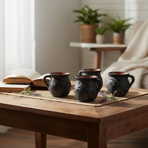 Puede incluir: Un juego de cuatro tazas pequeñas de cerámica negra redondeada con un interior de color terracota. Cada taza presenta un patrón de abanico texturizado y en relieve en la superficie exterior y un asa curva, dispuestas sobre una alfombrilla tejida.