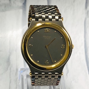 Könnte beinhalten: Eine Vintage-Armbanduhr mit dunkelgrauem Zifferblatt und goldfarbener Lünette. Die Uhr hat goldene Zeiger und Stundenmarkierungen. Das Armband ist eine Mischung aus silbernen und goldfarbenen Metallgliedern. Die Uhr wird auf einem transparenten Ständer ausgestellt.