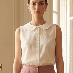 Op de afbeelding: Een mouwloze, crèmekleurige blouse met een ronde Peter Pan-kraag en een knoopsluiting aan de voorzijde, gecombineerd met een oudroze, getailleerde short met een hoge taille, plooien aan de voorzijde en een strakke zoom.