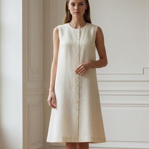 Op de afbeelding: Een mouwloze, knielange jurk in een effen off-white kleur. Het kledingstuk heeft een ronde halslijn en een verticale rij kleine, ronde knoopjes die in het midden aan de voorzijde naar beneden lopen. Het silhouet is recht en eenvoudig.