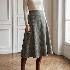 Puede incluir: Una mujer con un jersey de cuello alto color crema y una falda midi a cuadros grises. También lleva botas de cuero marrones. El conjunto es clásico y elegante, adecuado para diversas ocasiones.