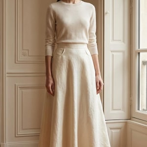 Op de afbeelding: Een vrouw draagt een crèmekleurige outfit, bestaande uit een trui met lange mouwen en een midi-rok met zakken. Ze draagt crèmekleurige hakken. De achtergrond is een kamer met een raam, wat een zachte, neutrale esthetiek creëert.
