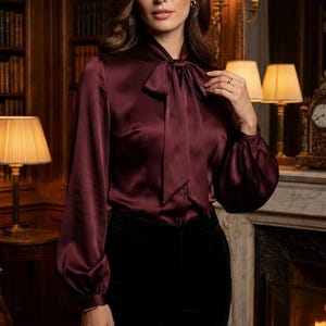 Op de afbeelding: Een bordeauxrode satijnen blouse met lange mouwen, een hoge hals met een gestrikte strik en knoopmanchetten, gecombineerd met een effen zwarte broek. De blouse heeft een gladde, glanzende afwerking en een gestructureerde pasvorm.