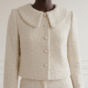 Può includere: Completo giacca e gonna in tweed color crema. La giacca presenta un colletto rotondo, bottoni perlati e maniche lunghe. Il tessuto ha un aspetto strutturato. L'outfit è esposto su un manichino, mostrando uno stile classico ed elegante.