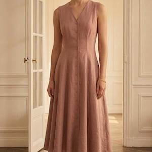 May include: A sleeveless midi dress in a dusty rose color featuring a V-neckline, a fitted bodice with vertical seam detailing, and a full skirt. The front of the dress is secured with a vertical row of matching buttons extending from the neckline.