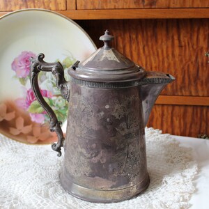 May include: An antique silver plated coffee pot with an ornate design and a lid. The pot is sitting on a white lace doily.
