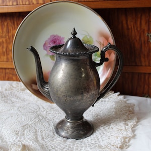 May include: A silver teapot with a decorative lid and handle. The teapot is sitting on a white doily.