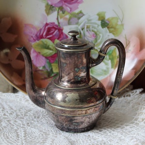 May include: A silver teapot with a gold trim and a floral design. The teapot has a round body and a curved spout. It is sitting on a white lace tablecloth.