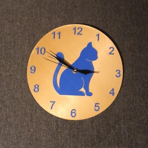 Könnte beinhalten: Eine runde Wanduhr mit hellbraunem Zifferblatt, blauen Ziffern und einer blauen Katzen-Silhouette. Die Uhrzeiger sind schwarz. Die Uhr ist ein dekorativer Artikel für ein Zuhause oder Büro.