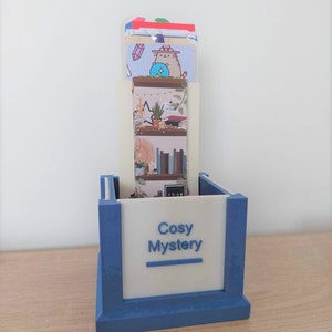 Könnte beinhalten: Ein blau-weißer Schreibtisch-Organizer mit dem Text "Cosy Mystery" auf der Vorderseite. Der Organizer enthält mehrere dekorative Karten mit Illustrationen einer Katze, Büchern und Pflanzen. Der Organizer steht auf einer Holzoberfläche.