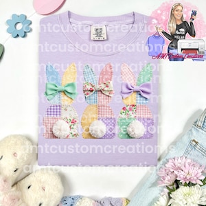 May include: A light purple Comfort Colors t-shirt featuring three Easter bunny designs. Each bunny has patchwork ears, a bow, and a fluffy white tail. The shirt has the text "mtcustomcreations" repeated across it.