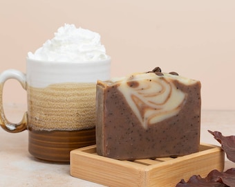 Puede incluir: Una pastilla de jabón artesanal con aroma a café y diseño en espiral, sobre un pequeño jabonero de madera. El jabón es marrón con acentos de granos de café. Una taza de cerámica llena de crema batida está al lado del jabón.