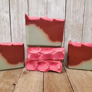 May include: Handmade soap bars in shades of red and green. The soap has a layered design with a wavy top. Some bars have a textured, floral-like pattern. The soaps are arranged on a wooden surface.