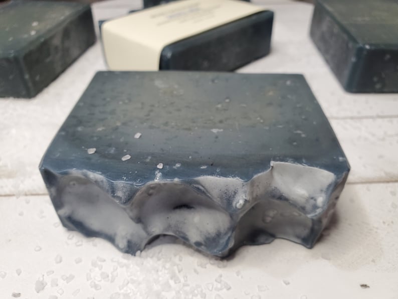 Peut inclure: Gros plan sur plusieurs pains de savon rectangulaires. Le savon est d'un gris bleu fonc&eacute; avec une surface textur&eacute;e et des accents blancs. Une barre a un bord rugueux et irr&eacute;gulier. Le savon semble avoir de petits cristaux blancs &agrave; la surface.