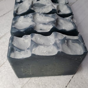 Peut inclure: Gros plan sur trois pains de savon faits &agrave; la main. Le savon a une base gris fonc&eacute; avec une couche sup&eacute;rieure blanche textur&eacute;e. La couche sup&eacute;rieure a un aspect marbr&eacute; avec de petites bosses irr&eacute;guli&egrave;res. Le savon est rectangulaire avec des bords l&eacute;g&egrave;rement arrondis.