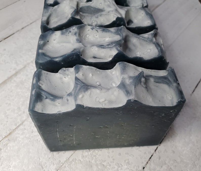Peut inclure: Gros plan sur trois pains de savon faits &agrave; la main. Le savon a une base gris fonc&eacute; avec une couche sup&eacute;rieure blanche textur&eacute;e. La couche sup&eacute;rieure a un aspect marbr&eacute; avec de petites bosses irr&eacute;guli&egrave;res. Le savon est rectangulaire avec des bords l&eacute;g&egrave;rement arrondis.