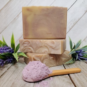 May include: Three bars of handmade soap stacked on top of each other. The soap is a light brown color with swirls of a darker brown color. A small wooden spoon with a scoop of pink clay powder is in the foreground. Lavender sprigs are on either side of the soap.