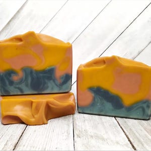 May include: Three rectangular bars of handcrafted soap featuring a marbled swirl pattern in shades of mustard yellow, muted peach, and dark slate blue. The soap bars have a smooth, matte finish with organic, flowing color transitions throughout.