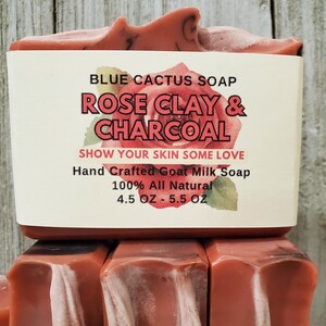 以下が含まれることがあります： 手作りのヤギミルク石鹸のスタック。一番上の石鹸には「BLUE CACTUS SOAP ROSE CLAY & CHARCOAL SHOW YOUR SKIN SOME LOVE Hand Crafted Goat Milk Soap 100% All Natural 127g - 156g」と書かれたラベルが付いています。石鹸は赤茶色です。