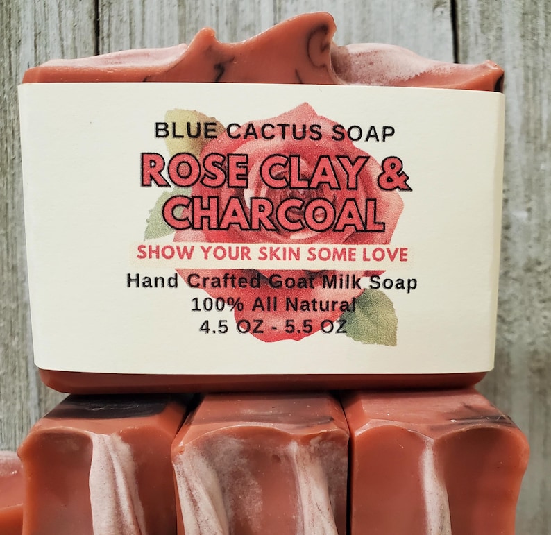 以下が含まれることがあります： 手作りのヤギミルク石鹸のスタック。一番上の石鹸には「BLUE CACTUS SOAP ROSE CLAY & CHARCOAL SHOW YOUR SKIN SOME LOVE Hand Crafted Goat Milk Soap 100% All Natural 127g - 156g」と書かれたラベルが付いています。石鹸は赤茶色です。