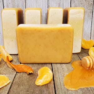May include: A collection of handmade soap bars in shades of yellow and orange, arranged on a wooden surface with mandarin orange slices, peels, and a honey dipper with honey. The soaps have a rectangular shape.