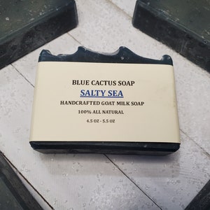 Peut inclure: Gros plan sur un pain de savon rectangulaire Blue Cactus, intitul&eacute; "Salty Sea". Le savon est fonc&eacute; avec une &eacute;tiquette claire. L'&eacute;tiquette indique "Handcrafted Goat Milk Soap" et "100% All Natural". Le pain de savon p&egrave;se entre 128 et 156 grammes.