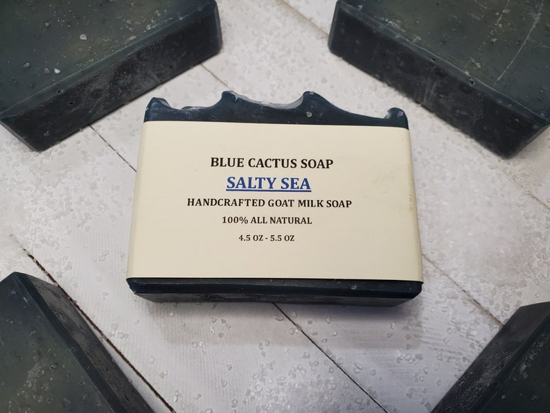Peut inclure: Gros plan sur un pain de savon rectangulaire Blue Cactus, intitul&eacute; "Salty Sea". Le savon est fonc&eacute; avec une &eacute;tiquette claire. L'&eacute;tiquette indique "Handcrafted Goat Milk Soap" et "100% All Natural". Le pain de savon p&egrave;se entre 128 et 156 grammes.