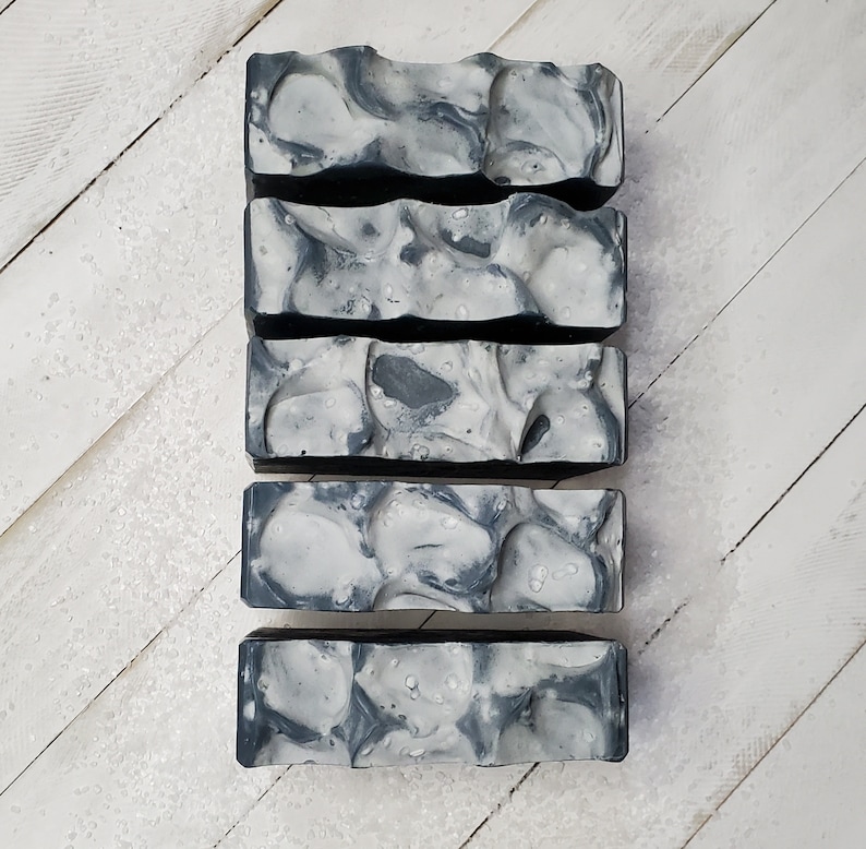 Peut inclure: Cinq pains de savon rectangulaires avec un motif marbr&eacute; gris et blanc. Les pains de savon ont une surface textur&eacute;e et in&eacute;gale, ressemblant &agrave; de la pierre. Le savon est dispos&eacute; verticalement sur une surface en bois blanc.