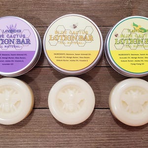 Op de afbeelding: Drie ronde blikken met Blue Cactus Lotion Bar, elk met een andere geur: Lavendel, Original en Yang Yang. De blikken zijn in een rij op een houten oppervlak gerangschikt. Elk blik heeft een wit etiket met zwarte tekst en een bijenillustratie. Onder de blikken bevinden zich drie witte lotion bars, elk met een bijenillustratie.