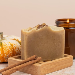 Könnte beinhalten: Herbstliche Anordnung mit einem dekorativen Kürbis, einem Seifenstück auf einer Holzseifenschale, Zimtstangen und einer brennenden Kerze in einem braunen Glas. Die Seife ist hellbraun mit strukturierter Oberfläche.