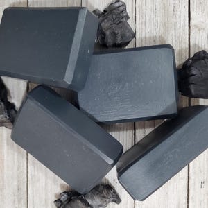 Könnte beinhalten: Vier schwarze Holzkohle-Seifenstücke auf einer Holzoberfläche. Die Seifenstücke sind rechteckig und haben eine glatte, matte Oberfläche.
