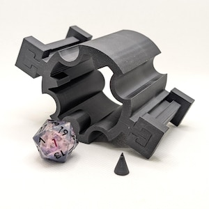 以下が含まれることがあります： 黒色の3Dプリントされたサイコロホルダー、多面体のサイコロ、小さな円錐形。 ホルダーは、幾何学的な切り欠きとサイコロ用のスロットを備えた湾曲したデザインです。 サイコロは透明で、ピンクと紫の色合いがあり、数字が見えます。