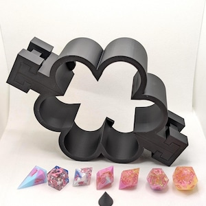 May include: A black, 3D-printed dice tray shaped like a four-leaf clover, with a set of colourful polyhedral dice. The dice are various colours, including pink, blue, and clear with glitter. A small black cone is also present.