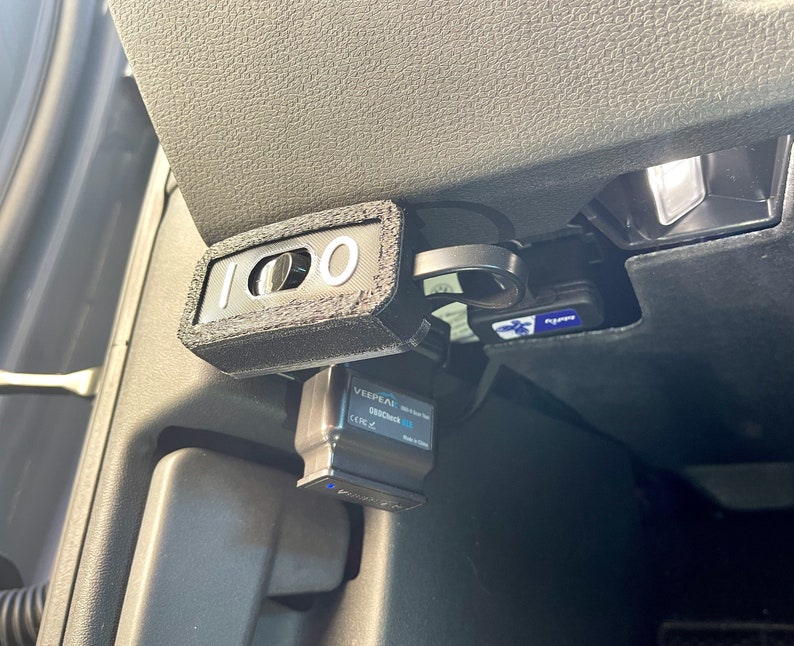 May include: A black OBD2 adapter with the text "VEEPEAK OBDCheck BT" and a black device with the number "100" printed on it. The device is connected to a car's diagnostic port.