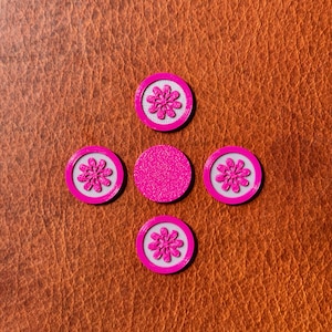 Puede incluir: Cinco círculos de papel redondos de color rosa y blanco con un diseño de flor rosa en el centro. Los círculos están dispuestos en forma de cruz sobre un fondo de cuero marrón.