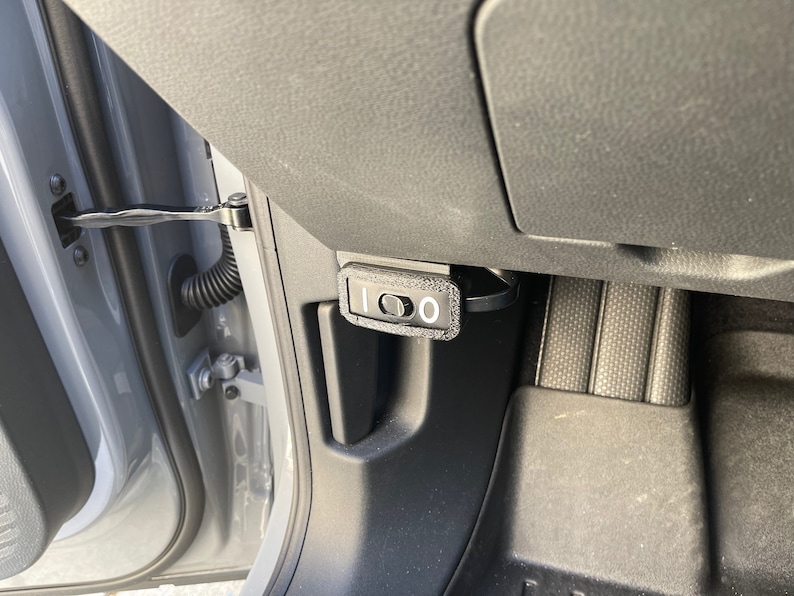 May include: A black plastic switch with a white label that says "1 0" is mounted on the side of a car's interior. The switch is located near the door hinge.