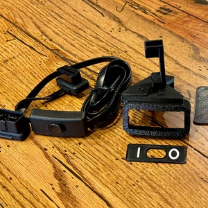 May include: A black automotive diagnostic connector with a black switch and a black cable. There are also three black plastic parts, one with a rectangular opening and two with a rectangular shape.