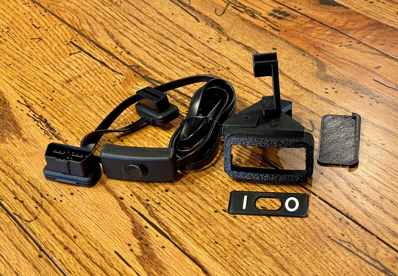 May include: A black automotive diagnostic connector with a black switch and a black cable. There are also three black plastic parts, one with a rectangular opening and two with a rectangular shape.