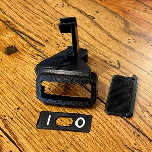 May include: A black 3D printed phone stand with a slot for a phone and a separate black 3D printed piece with the number "100" printed in white.