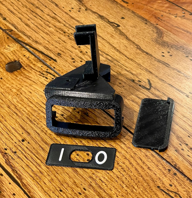 May include: A black 3D printed phone stand with a slot for a phone and a separate black 3D printed piece with the number "100" printed in white.