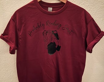 May include: A maroon t-shirt with a black graphic of a woman reading a book. The text "probably reading" and "shirt" are written in black cursive above and below the graphic.