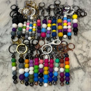 May include: A collection of beaded keychains in various colors, including purple, pink, blue, yellow, and green. Each keychain features a series of round beads strung together, with metal key rings and clasps.