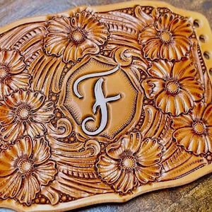 Może przedstawiać: Ręcznie wykonany skórzany element z kwiatowym wzorem i literą "F" pośrodku. Brązowa skóra jest misternie rzeźbiona w kwiaty i liście, a krawędzie mają równomiernie rozmieszczone otwory. Całość jest symetryczna i szczegółowa.