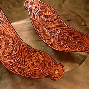 May include: Two brown leather straps with intricate floral designs, possibly for a cowboy boot or other leather goods.