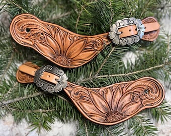 May include: A pair of brown leather spur straps with intricate floral designs and silver buckles. The straps are decorated with a sunflower motif.