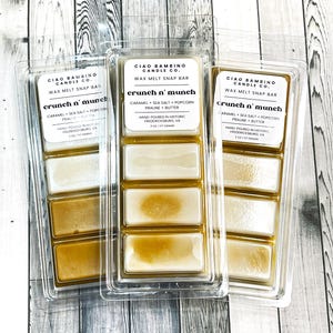 May include: Three clear plastic containers with three wax melt snap bars in each. The wax melts are labeled "Ciao Bambino Candle Co. Crunch n' Munch" and list the ingredients as caramel, sea salt, popcorn, praline, and butter. The wax melts are hand-poured in historic Fredericksburg, VA.
