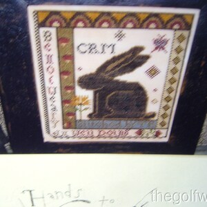 May include: A framed cross-stitch design featuring a black rabbit with the text "Be Not Afraid" and "God Is Love" on the left and right sides of the rabbit, and the text "Celi" at the top. The background is a white fabric with a brown, red, and yellow patterned border.