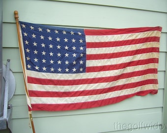 Può includere: Una bandiera americana vintage con 48 stelle, con strisce rosse e bianche e un campo blu con stelle bianche. La bandiera è montata su un'asta di legno ed è esposta all'aperto.