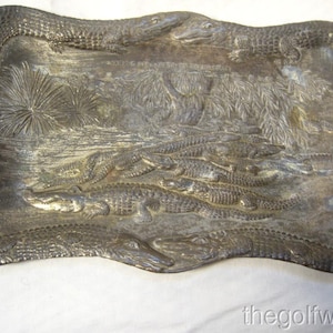 May include: Silver-coloured rectangular tray featuring a detailed relief of alligators in a swamp scene. The tray has a wavy border, and the alligators are shown in various sizes and poses, creating a textured surface.